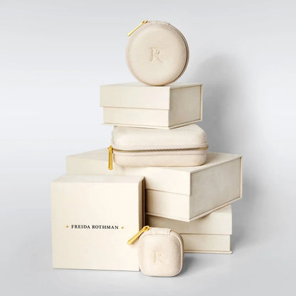 An artfully arranged stack of elegant beige jewelry boxes of various sizes, each featuring gold zipper accents. The largest box at the bottom showcases &quot;FREIDA ROTHMAN&quot; imprinted in 14K matte gold. Inside, nestled safely, is a Midnight Tri-Tone Twilight Oval Station Tennis Bracelet adorned with cubic zirconia stones from the brand Midnight.