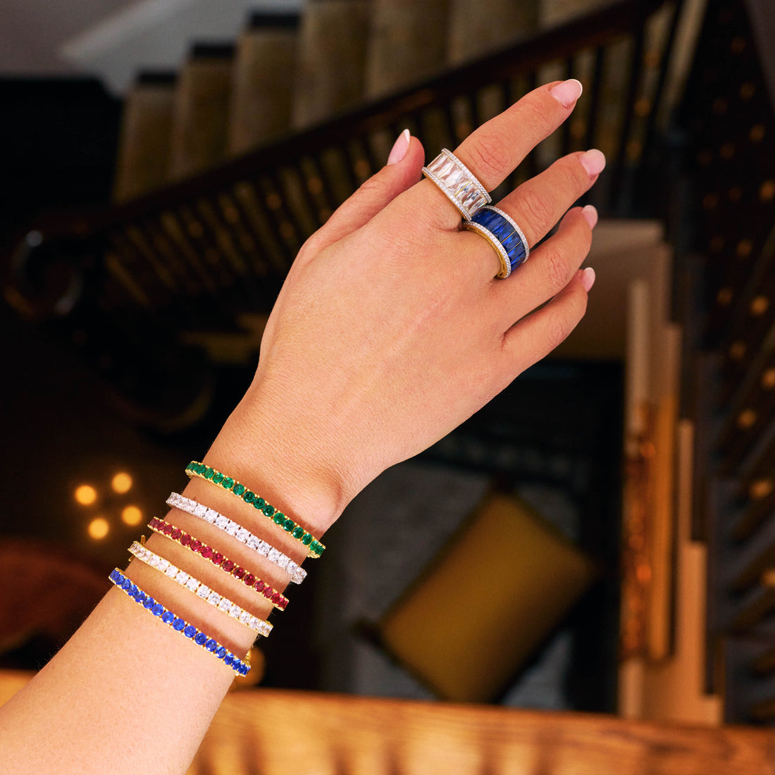 A hand adorned with several colorful beaded bracelets and rings is extended over a staircase with a dark wooden railing. Among the bracelets, the Midnight Tri-Tone Twilight Tennis Bracelet by Midnight stands out with its elegant mix of green, red, blue, and gold beads. The rings feature cubic zirconia stones and intricate designs.