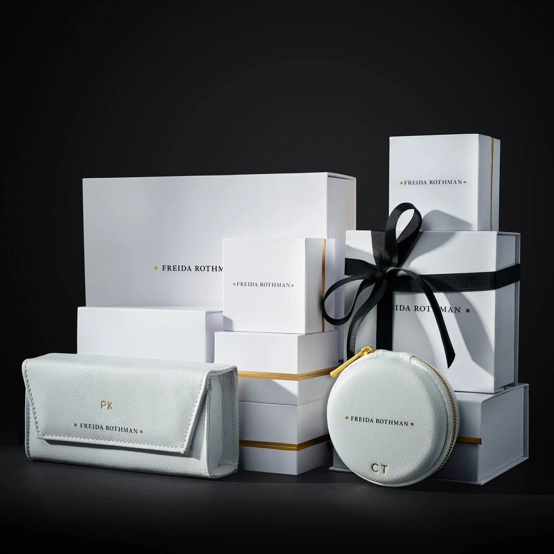 A stack of white gift boxes with gold-trimmed edges and a subtle rhodium matte finish, elegantly labeled &quot;FREIDA ROTHMAN,&quot; against a dark background. In front, there&